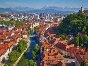 A view of a Slovenian town with orange roofs and bright green trees popping up in between, with mountains off in the distance.